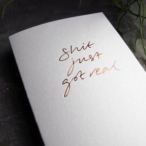Close up of luxury white greetings card with "Shit Just Got Real" handwritten in the front and hand printed in rose gold foil on a grey background with some green plant leaves at the side.