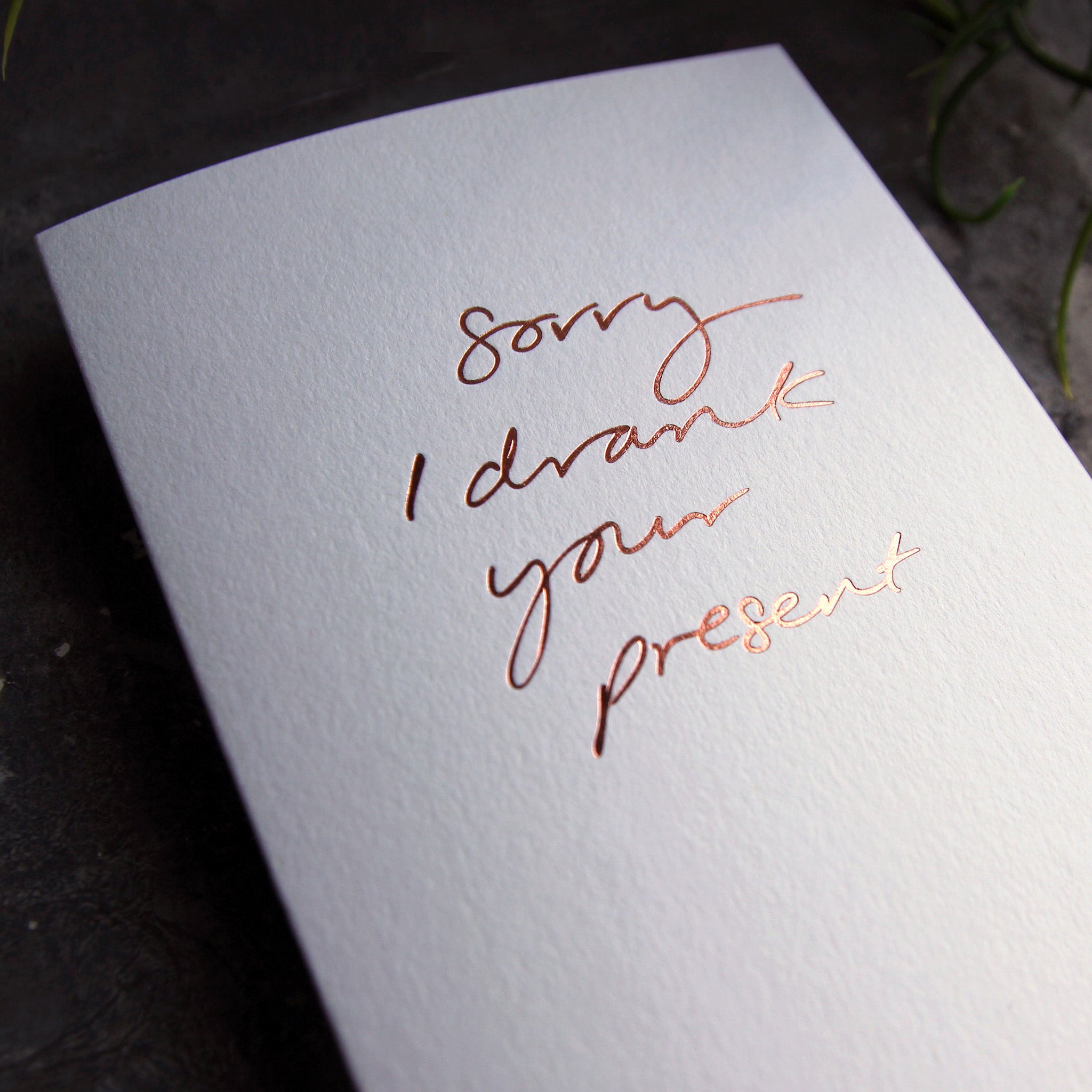 Close up of luxury white greetings card with "Sorry I Drank Your Present" handwritten in the front and hand printed in rose gold foil on a grey background with some green plant leaves at the side.