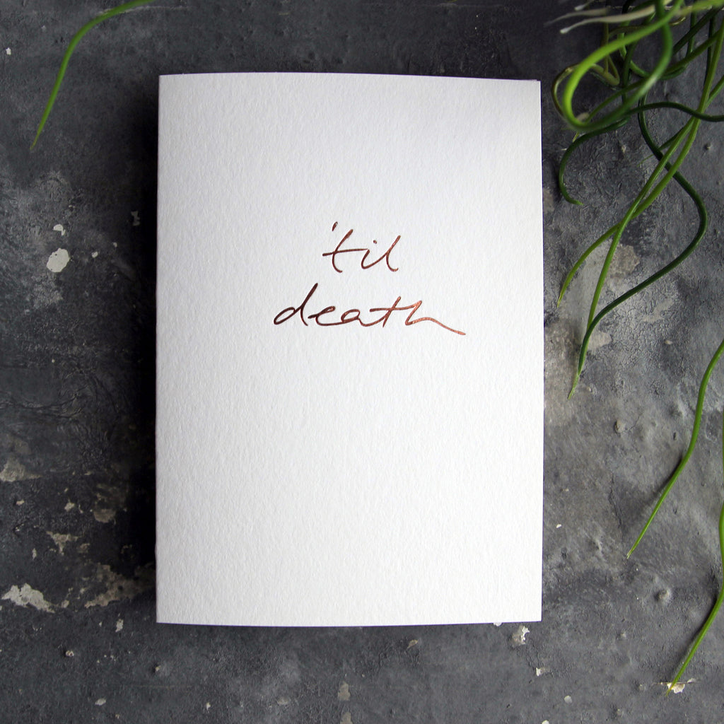 Luxury white greetings card with "Til Death" handwritten in the front and hand printed in rose gold foil on a grey background with some green plant leaves at the side.