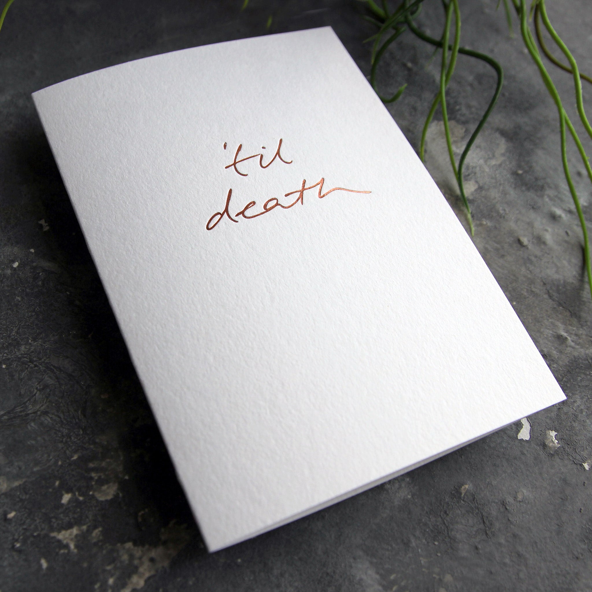 Luxury white greetings card with "Til Death" handwritten in the front and hand printed in rose gold foil on a grey background with some green plant leaves at the side.