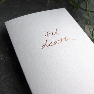 Close up of luxury white greetings card with "Til Death" handwritten in the front and hand printed in rose gold foil on a grey background with some green plant leaves at the side.