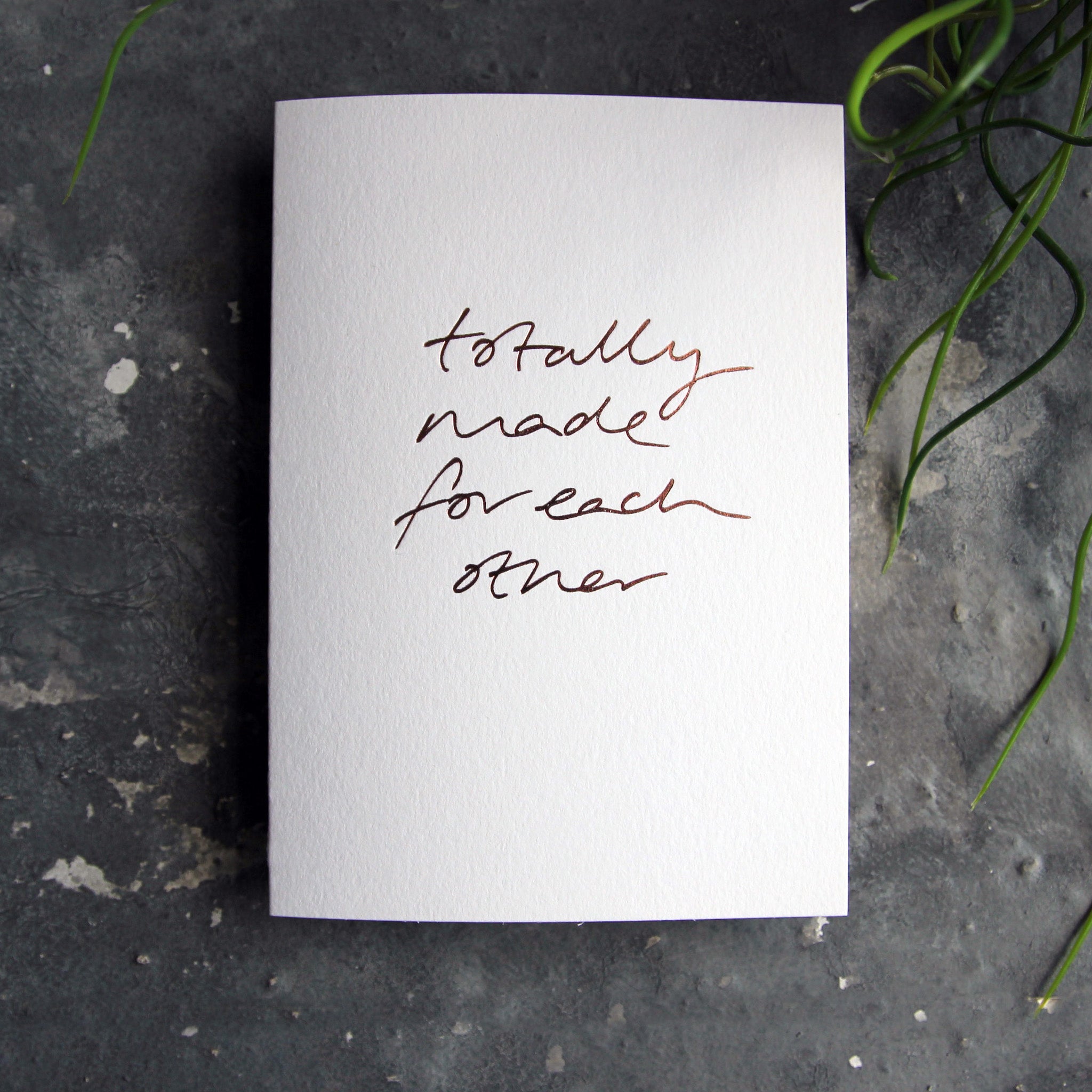 Luxury white greetings card with "Totally Made For Each Other' handwritten in the front and hand printed in rose gold foil on a grey background with some green plant leaves at the side.