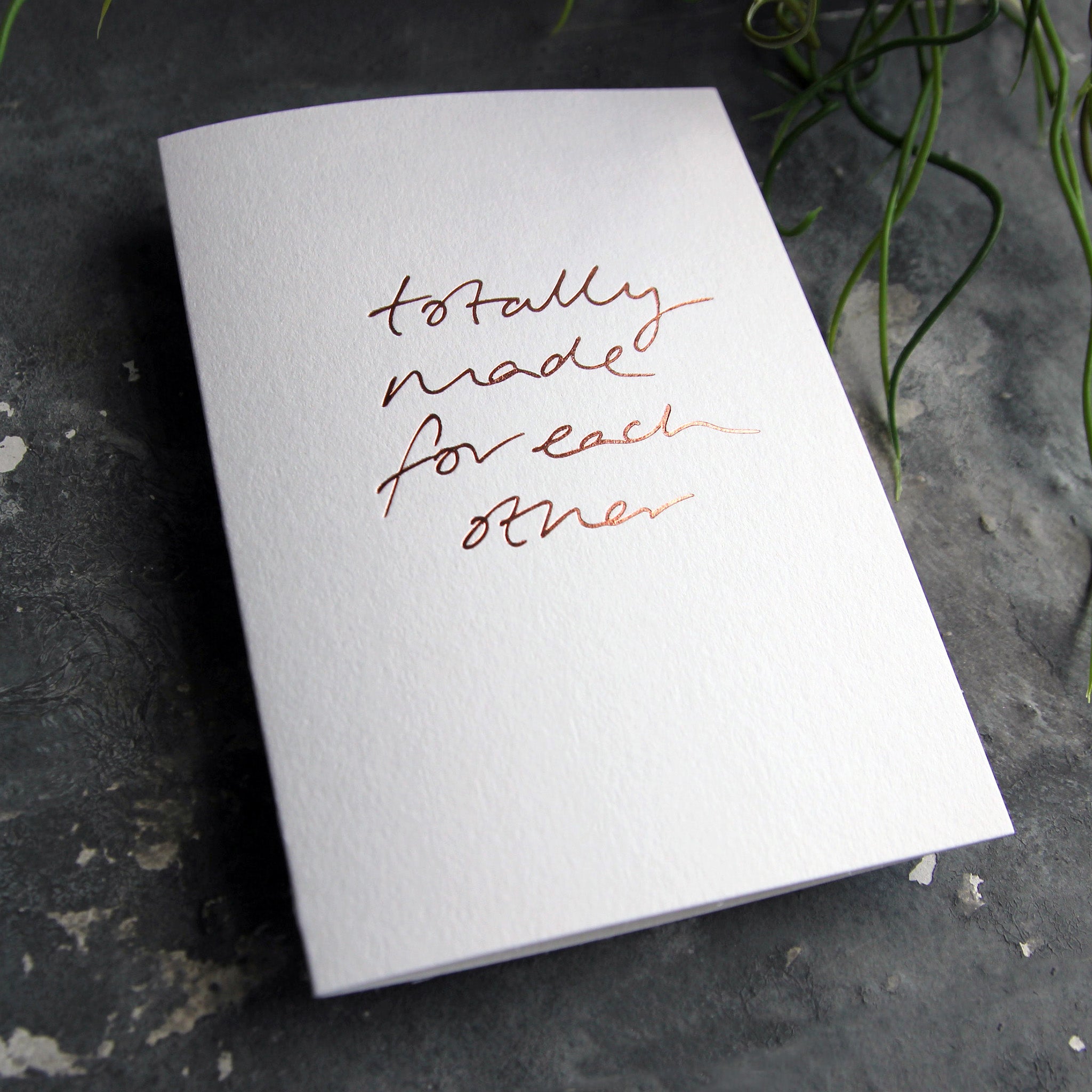 Luxury white greetings card with "Totally Made For Each Other' handwritten in the front and hand printed in rose gold foil on a grey background with some green plant leaves at the side.