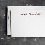 Luxury white notecard and envelope with "What The Hell" handwritten on the front and hand printed in rose gold foil on a grey background.