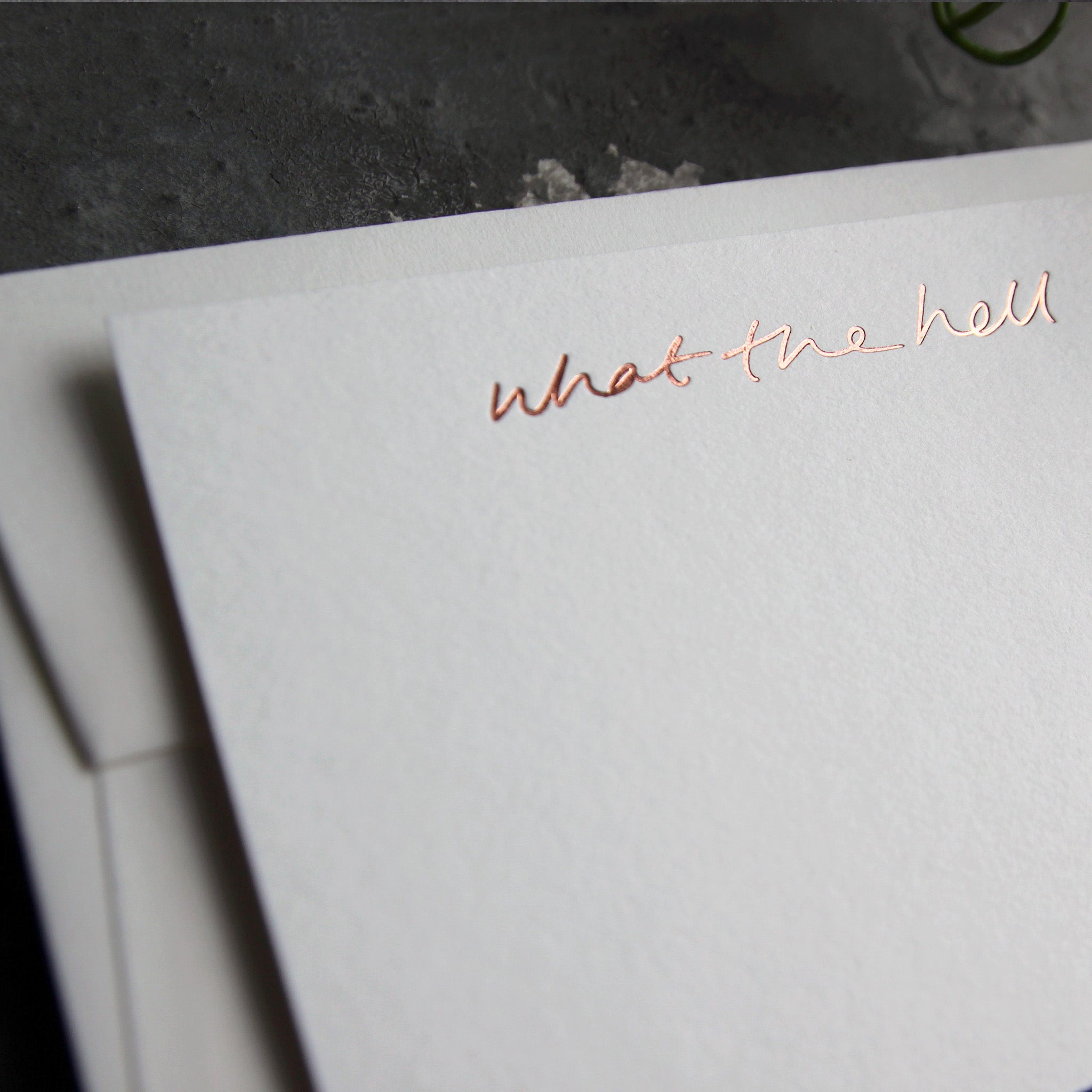 Close up of luxury white notecard and envelope with "What The Hell" handwritten on the front and hand printed in rose gold foil on a grey background.