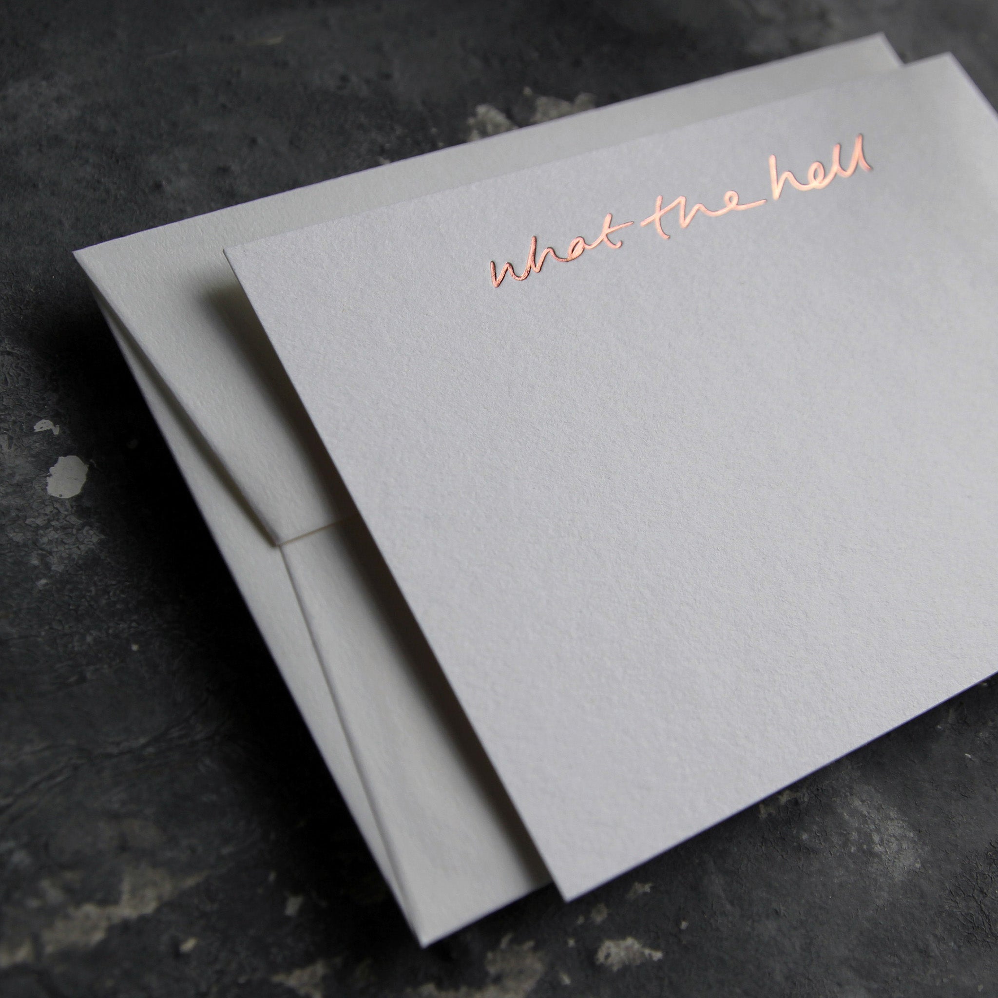 Luxury white notecard and envelope with "What The Hell" handwritten on the front and hand printed in rose gold foil on a grey background.