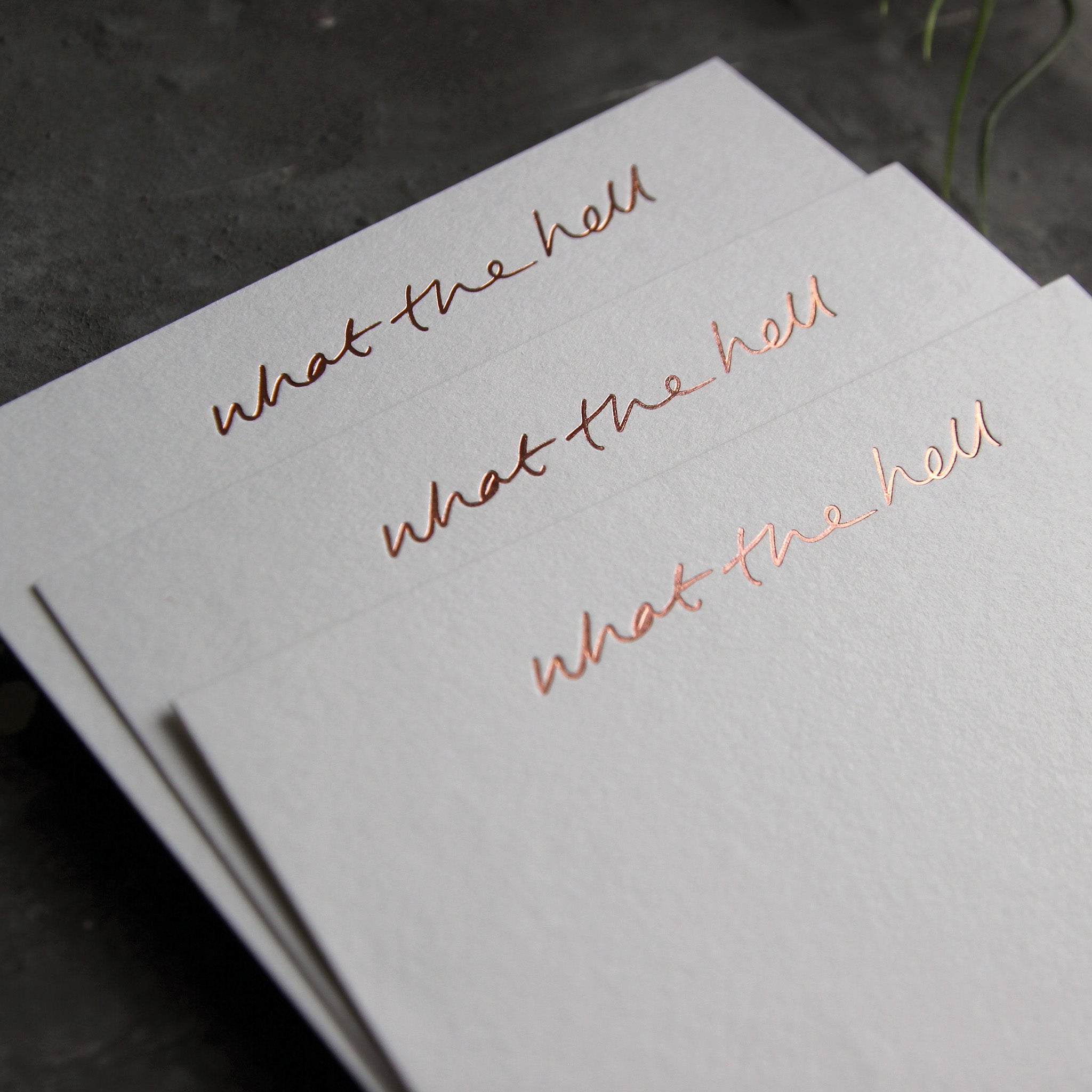 Close up of 3 luxury white notecards with "What The Hell" handwritten on the front and hand printed in rose gold foil on a grey background.