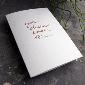 Luxury white greetings card with "You Deserve Each Other' handwritten in the front and hand printed in rose gold foil on a grey background with some green plant leaves at the side.