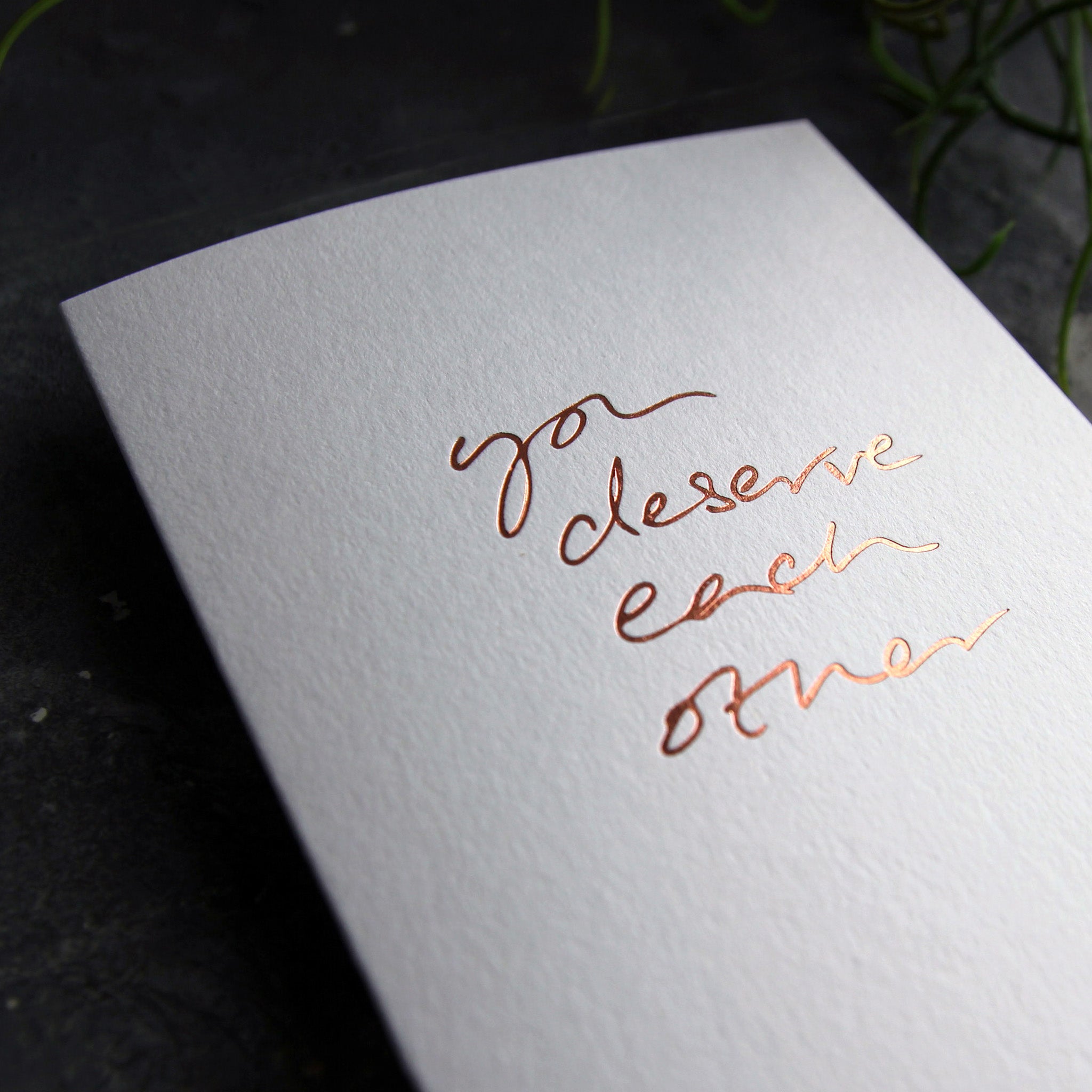 Close up of luxury white greetings card with "You Deserve Each Other' handwritten in the front and hand printed in rose gold foil on a grey background with some green plant leaves at the side.