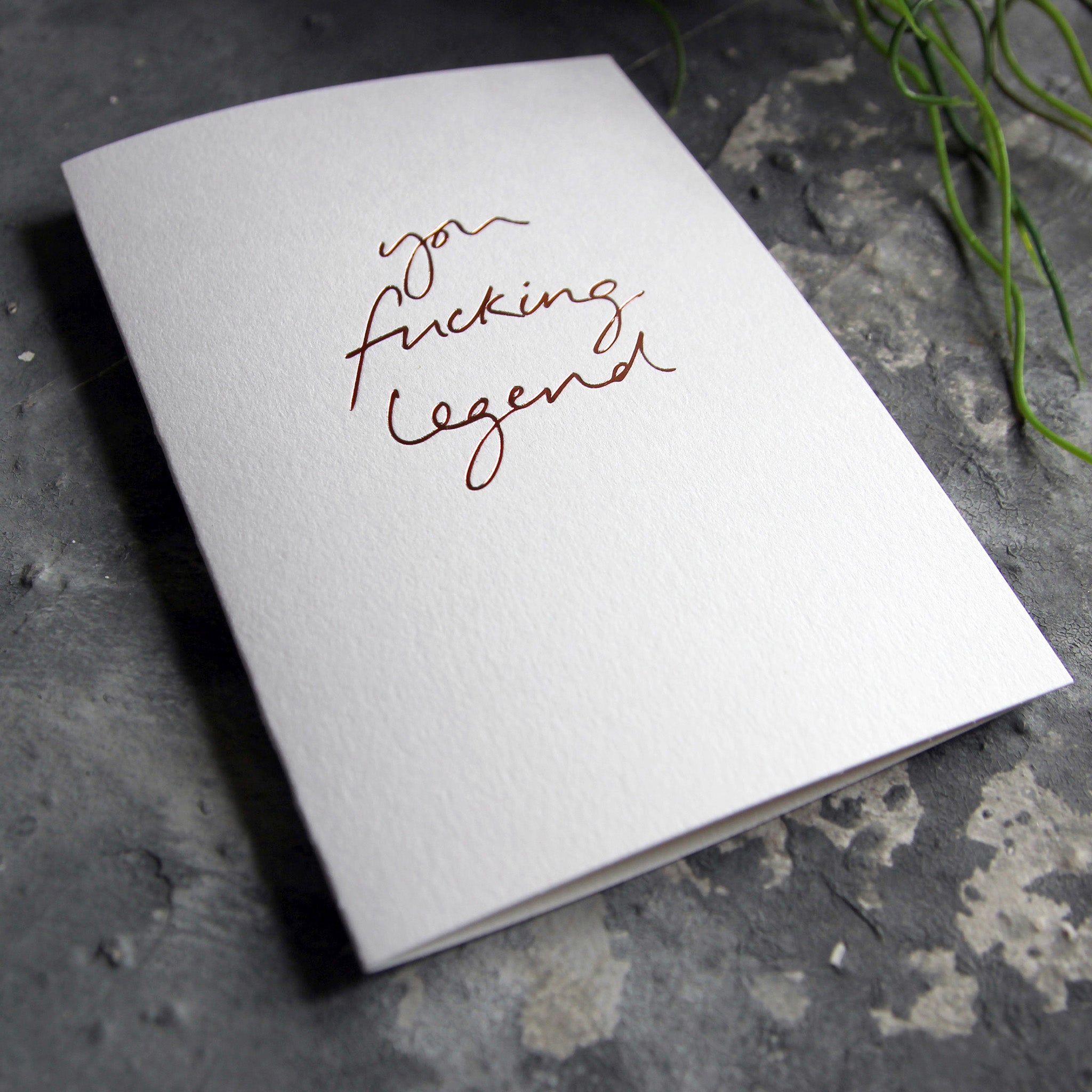 Luxury white greetings card with "You Fucking Legend' handwritten in the front and hand printed in rose gold foil on a grey background with some green plant leaves at the side.