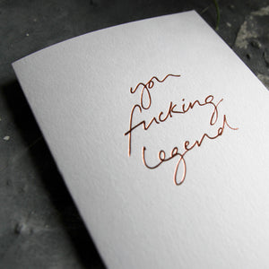 Close up of luxury white greetings card with "You Fucking Legend' handwritten in the front and hand printed in rose gold foil on a grey background with some green plant leaves at the side.