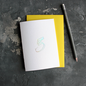 A fifth birthday card with a hand drawn number five hand pressed in holographic foil on the front and a yellow envelope