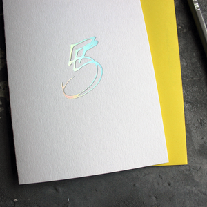 A fifth birthday card with a hand drawn number five hand pressed in holographic foil on the front and a yellow envelope