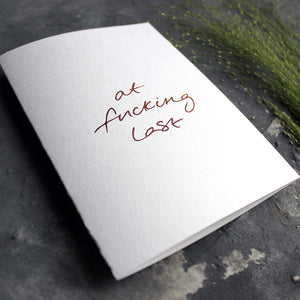 this hand foiled card says 'at fucking last' on the front in handwriting on white coloured paper