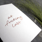 this hand foiled card says 'at fucking last' on the front in handwriting on white coloured paper