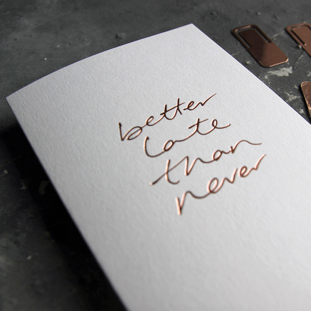 This birthday card says Better Late Than Never is handwritten design and handfoiled in rose gold foil on luxury white paper, photographed on a grey board 
