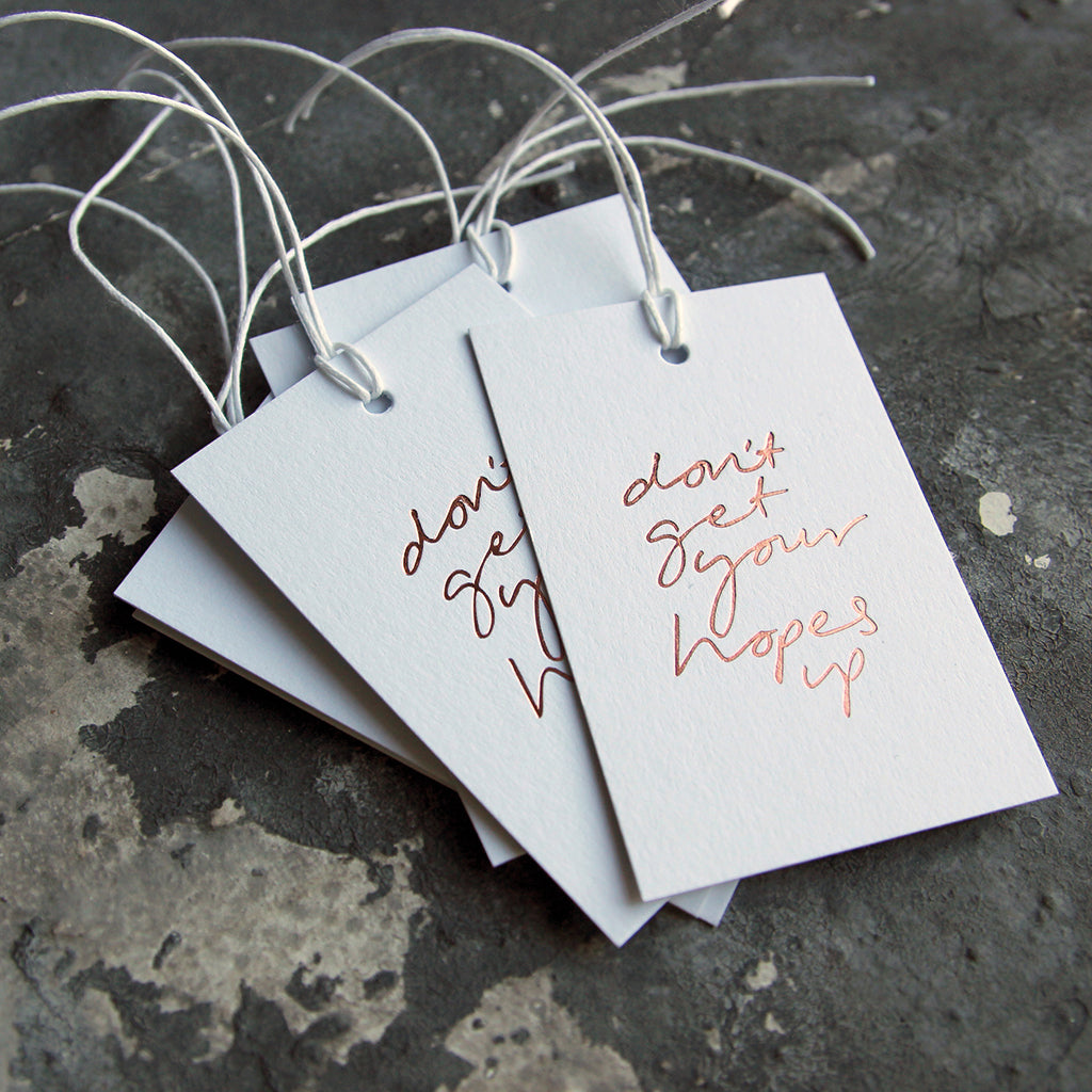 This cash card saPack of gift tags say Don't Get Your Hopes Up handwritten and hand printed in rose gold foil on white luxury paper on grey board