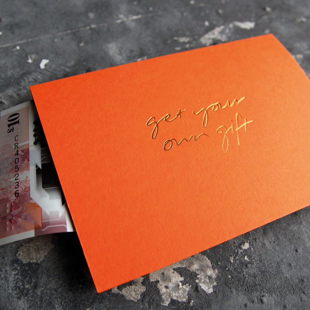 This cash card says Get Your Own Gift and is handwritten and hand printed in gold foil on orange luxury paper on grey board