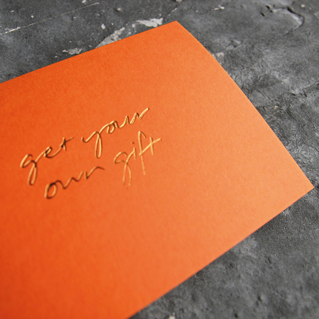 This cash card says Get Your Own Gift and is handwritten and hand printed in gold foil on orange luxury paper on grey board