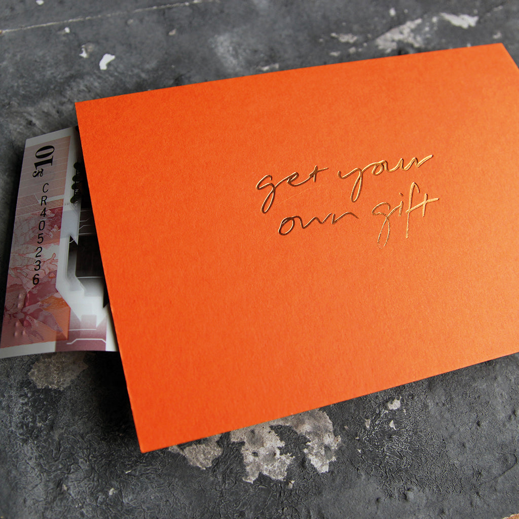 This cash card says Get Your Own Gift and is handwritten and hand printed in gold foil on orange luxury paper on grey board