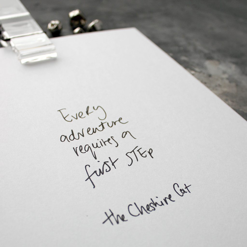 This handwritten children's cheshire cat print says 'Every Adventure Requires A First Step' in black on white paper. 