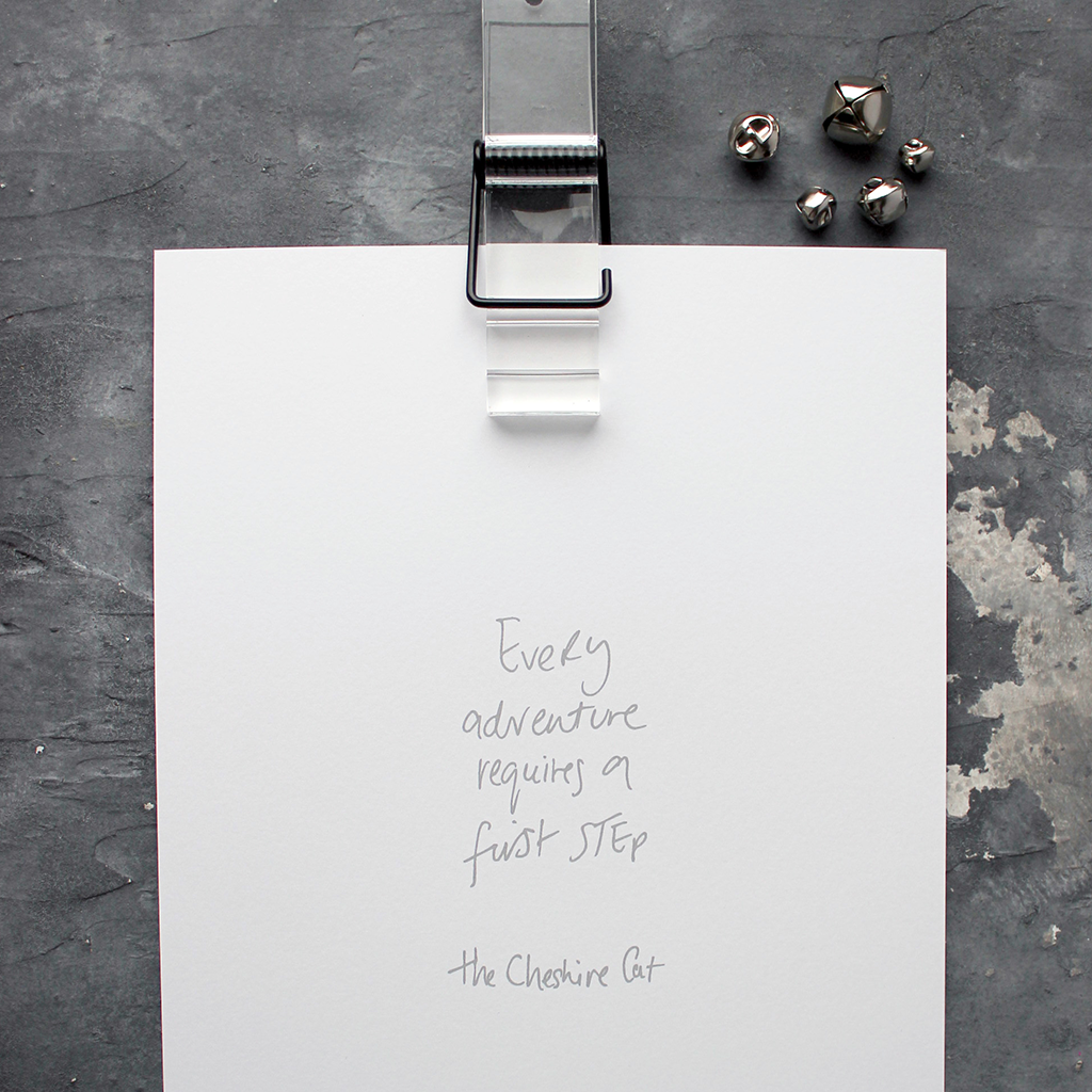 This handwritten children's cheshire cat print says 'Every Adventure Requires A First Step' in grey on white paper. 
