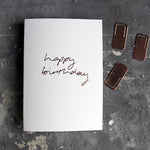 This birthday card says Happy Birthday is handwritten design and handfoiled in rose gold foil on luxury white paper, photographed on a grey board 
