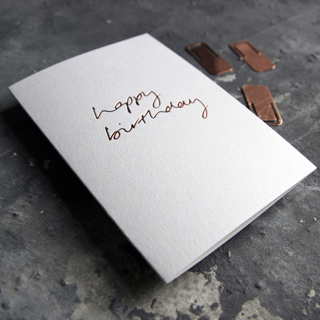 This birthday card says Happy Birthday is handwritten design and handfoiled in rose gold foil on luxury white paper, photographed on a grey board 
