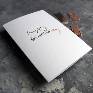 This birthday card says Happy Birthday is handwritten design and handfoiled in rose gold foil on luxury white paper, photographed on a grey board 