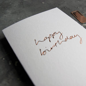 This birthday card says Happy Birthday is handwritten design and handfoiled in rose gold foil on luxury white paper, photographed on a grey board 