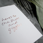 The front of the card has the phrase 'here's to the shits and giggles' handwritten and hand pressed in rose gold foil