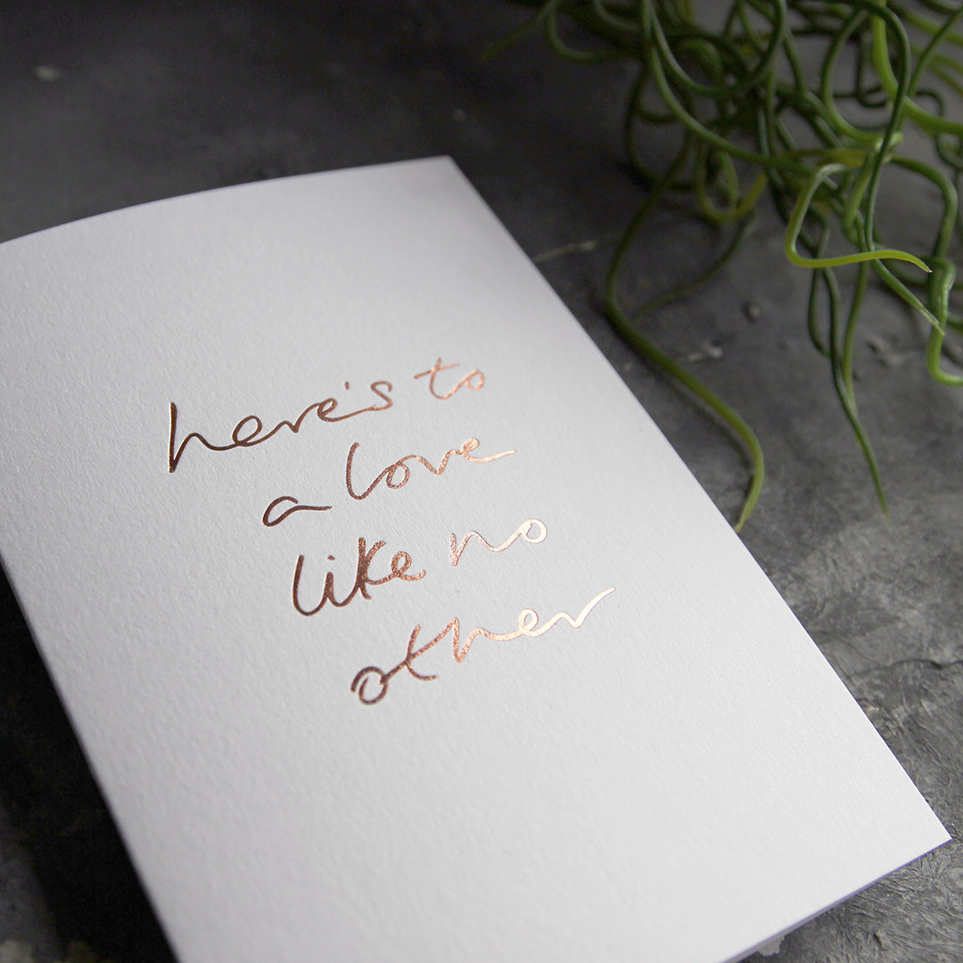 this hand foiled luxury white card says Here's To A Love Like No Other on the front in rose gold foil