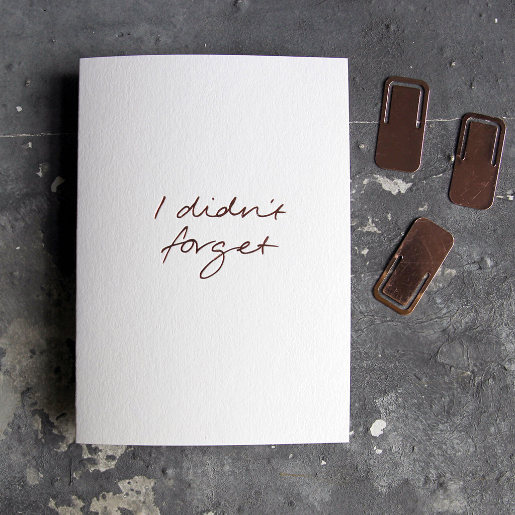 This birthday card says I Didn't Forget is handwritten design and handfoiled in rose gold foil on luxury white paper, photographed on a grey board 