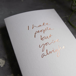 this hand foiled luxury white card says I Hate People But You're Alright on the front in rose gold foil