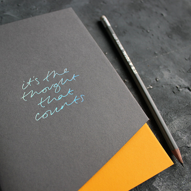 a grey and yellow coloured handmade luxury notebook that's hand foiled with the message It's The Thought That Counts in holographic foil
