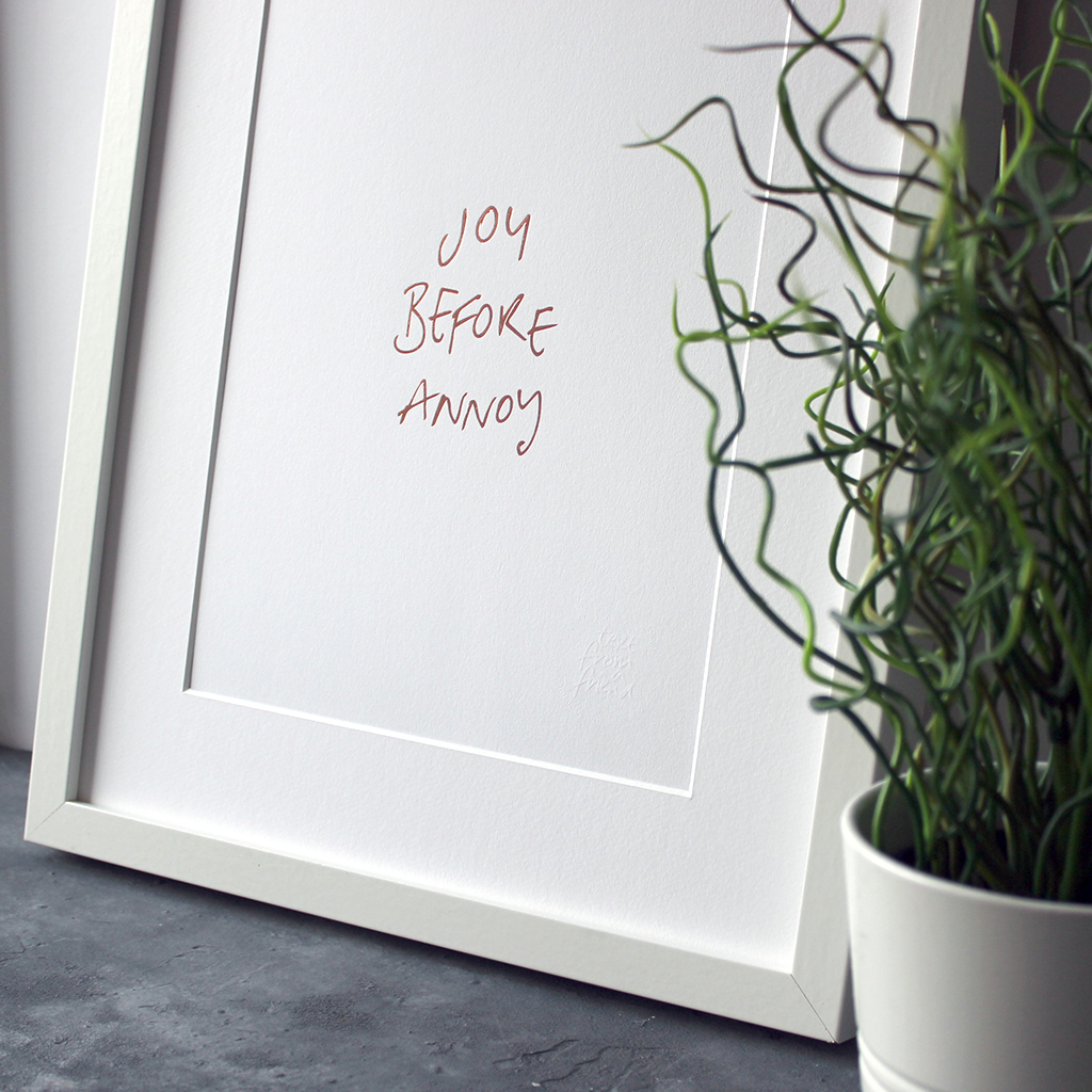 This unframed rose gold foil print has the words Joy before Annoy handwritten in a contemporary design