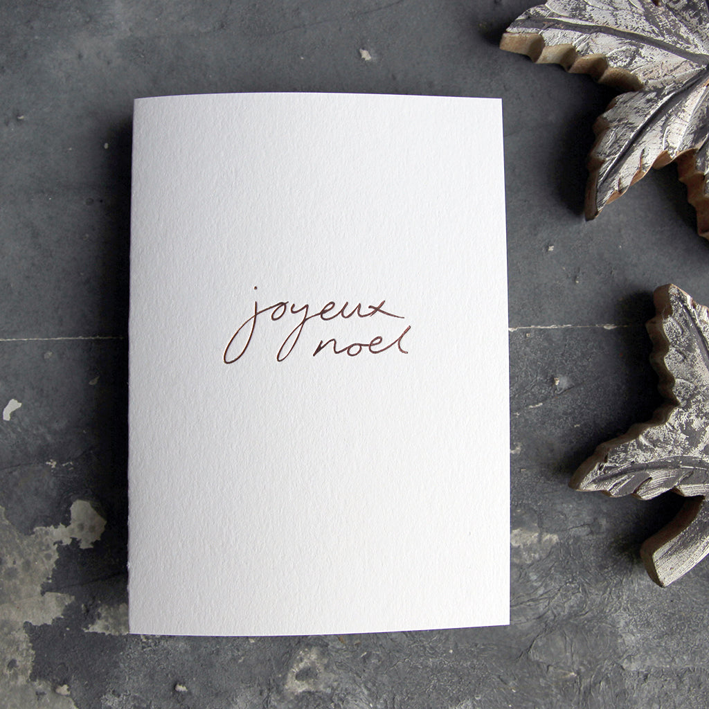 A Joyeux Noel christmas card on a grey board is handwritten and handfoiled on a luxury white paper