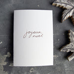 A Joyeux Noel christmas card on a grey board is handwritten and handfoiled on a luxury white paper