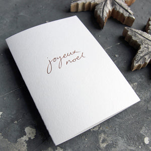 A Joyeux Noel christmas card on a grey board is handwritten and handfoiled on a luxury white paper
