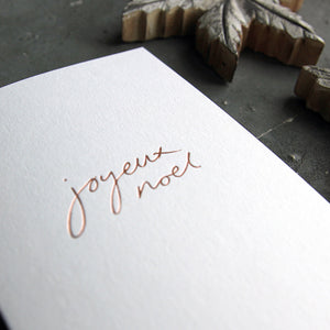 A Joyeux Noel christmas card on a grey board is handwritten and handfoiled on a luxury white paper