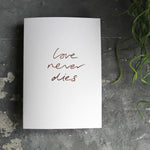 this hand foiled luxury white card says Love Never Dies on the front in rose gold foil