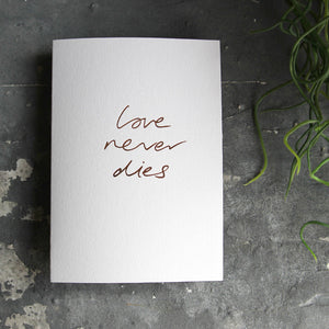 this hand foiled luxury white card says Love Never Dies on the front in rose gold foil