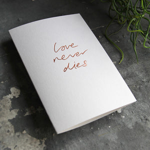 this hand foiled luxury white card says Love Never Dies on the front in rose gold foil