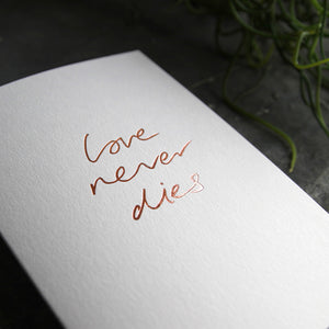 this hand foiled luxury white card says Love Never Dies on the front in rose gold foil