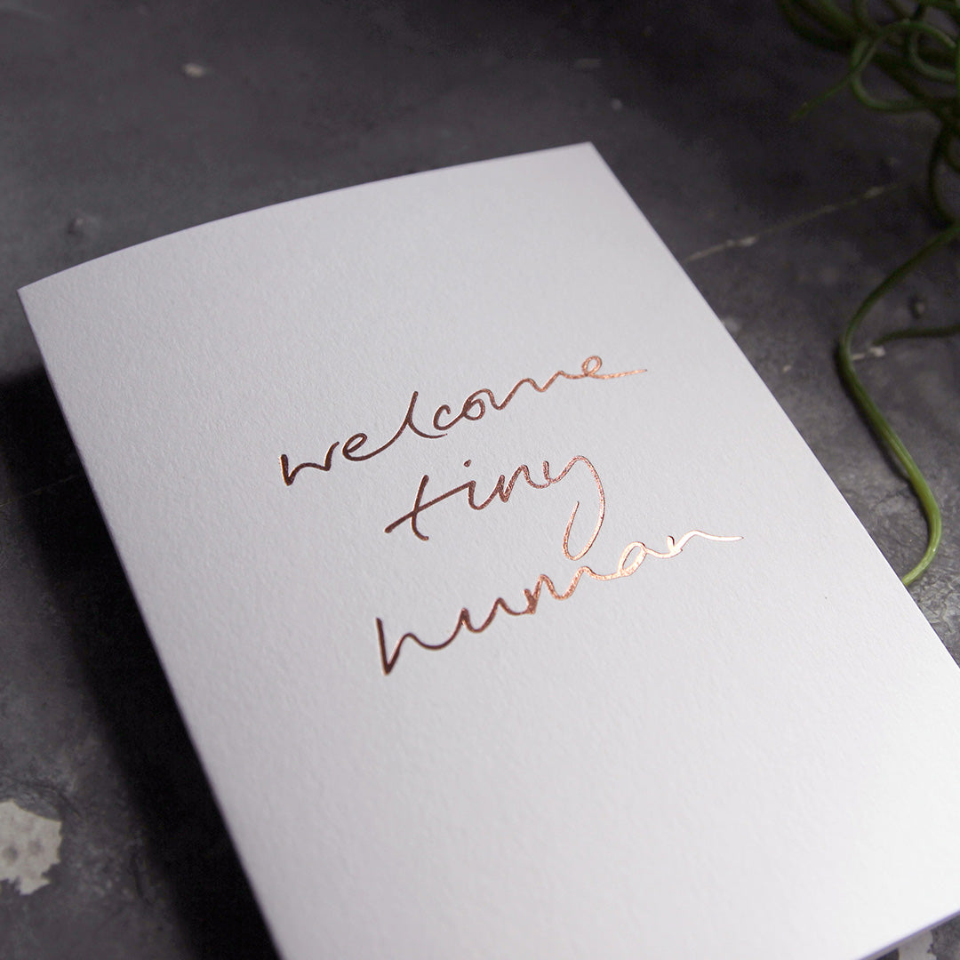 this hand foiled luxury white card says Welcome Tiny Human on the front in rose gold foil