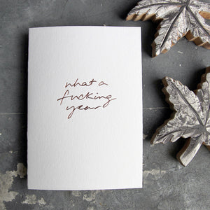 This christmas card says What A Fucking Year on hand foiled on white luxury paper on grey board