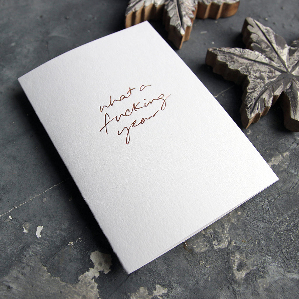 This christmas card says What A Fucking Year on hand foiled on white luxury paper on grey board