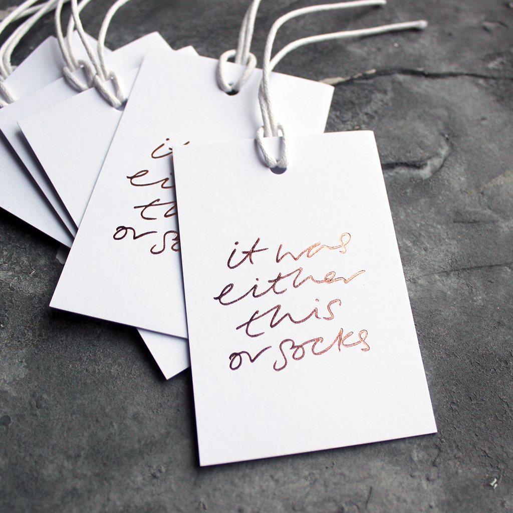 Luxury white gift tags with waxed cotton thread have "It Was Either This Or Socks' handprinted in handwritten rose gold foil.