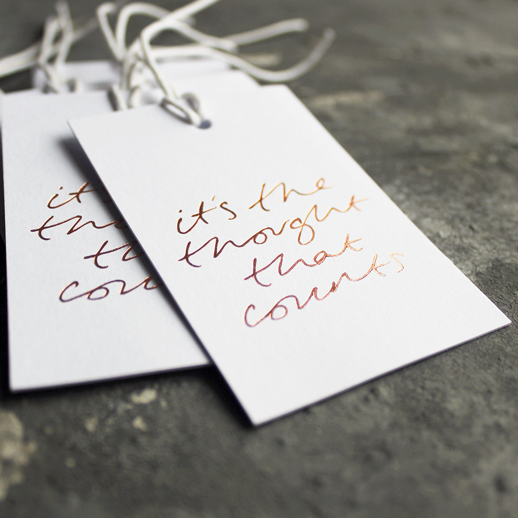 Luxury white gift tags with waxed cotton thread have "It's The Thought That Counts' handprinted in handwritten rose gold foil.