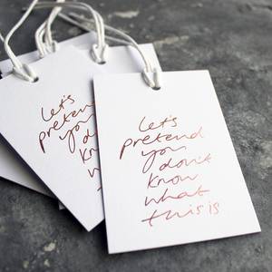 Luxury white gift tags with waxed cotton thread have "Let's Pretend You Don't Know What This Is' handprinted in handwritten rose gold foil.
